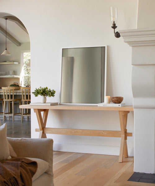 A light wood console table holds a framed mirror, the Untitled 5 Framed Print, a plant, a book, a candle, and a bowl against a white wall. An archway opens to a dining area with table and chairs in the background.
