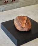 A loaf of rustic, crusty bread sits on a Square Butcher Block atop a light-colored kitchen counter with cabinets and a stove in the background.