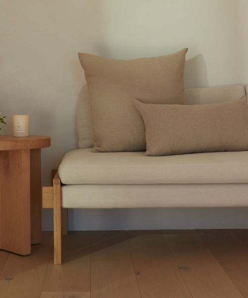 A minimalist living space features a beige sofa with two Linen Pillows made from natural linen and feather down inserts, set on a light wood floor beside a wooden side table with a lit candle for a calm, cozy vibe.