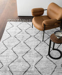 A modern tan armchair and small round side table rest on the Nala Rug, featuring a natural wool base with a black zigzag pattern and fringe, set atop a wooden floor near a white wall.