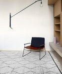 A minimalist room features a modern chair with a black leather seat, the hand-knotted Nala Rug in natural wool, built-in wooden shelves, a pink vase, and a long-arm wall lamp with an exposed bulb.