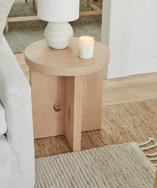 A light wood side table by a beige sofa holds a round white lamp and a lit candle. The table stands on the tan woven sustainable River Rug, with part of a cream textured rug seen in the foreground.