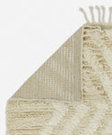 The Cabin Rug from Jenni Kayne Rugs is a cream-colored, textured piece with a geometric pattern, flipped corner showing its woven underside and fringe—a Moroccan-inspired rug that adds timeless elegance to any space.