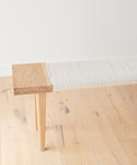 The Oak Day Bench features a minimalist design with hand-wrapped rope, solid wooden legs, and a light-colored woven seat, set on a light wood floor against a white wall.