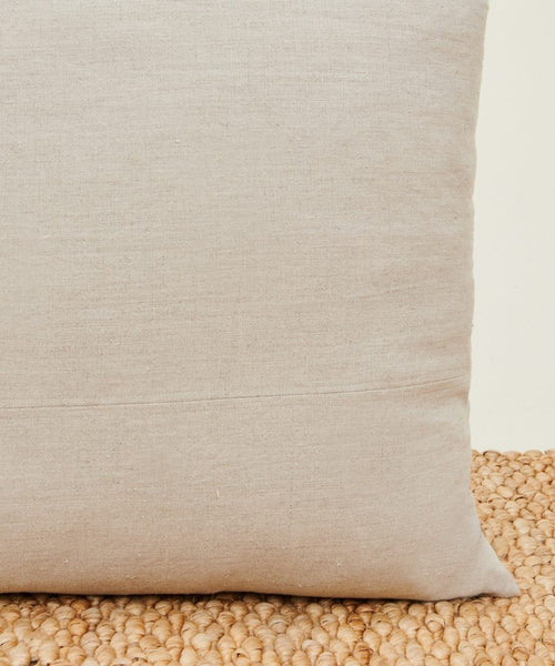 A close-up of the Linen Pillow in light beige natural linen with a smooth texture and feather down insert, displayed on a woven, natural-colored mat against an off-white background for a soft, neutral look.