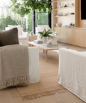 Bright modern living room with white armchairs, neutral decor, a Rug Swatch jute rug, wooden coffee table with books and a plant, large windows, and built-in wooden shelves displaying decorative items.