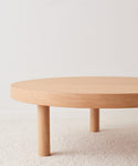 The Bay Coffee Table, crafted from knotty alder, showcases a minimalist round form with three cylindrical legs and is displayed on a light textured carpet in front of a plain white wall.