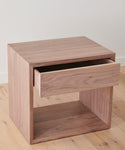The Berkeley Nightstand, crafted from solid walnut, features clean lines, a single open drawer, and a natural wood finish. It is handmade and shown on a light wood floor against a plain white wall.