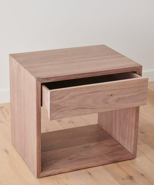 The Berkeley Nightstand, crafted from solid walnut, features clean lines, a single open drawer, and a natural wood finish. It is handmade and shown on a light wood floor against a plain white wall.