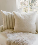 An Ojai Pillow and a solid pillow sit on a white sofa. A cozy, light faux fur throw lies in front, while a window with tall grass can be seen in the background.