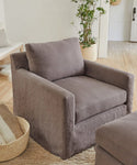 A modern living room featuring a plush, charcoal armchair in Fabric Swatch Charcoal Linen on a woven rug, a matching ottoman, a basket with blankets, a potted plant, and light wood floors.