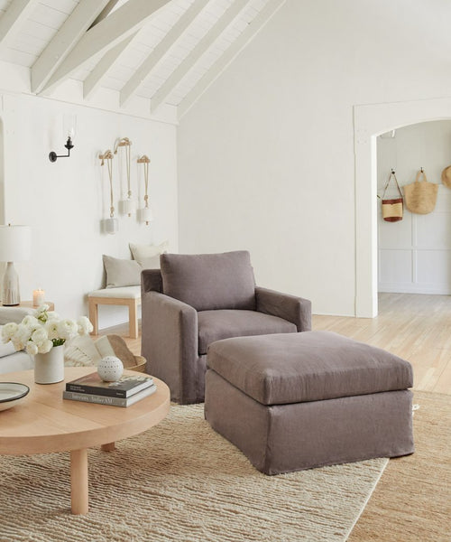 A cozy living room features the Harbor Chair, a charcoal linen chair handmade in Los Angeles, with a matching ottoman on a beige rug. Nearby, a round wooden coffee table displays books, flowers, and candles atop wood floors and light walls.