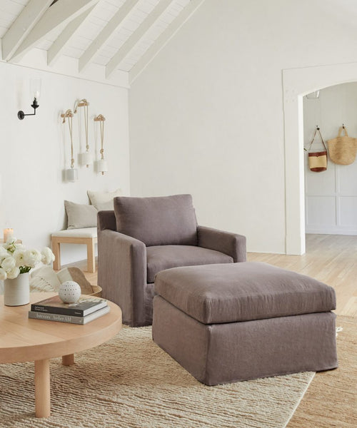 A cozy living room features a gray armchair and the Harbor Ottoman in charcoal linen, a wooden coffee table with books and decor, light wood floors, white walls with minimalist decorations, and hanging baskets.
