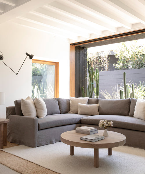 A bright, modern living room features a spacious Harbor Sectional in Belgian Flax Linen, a round wooden coffee table with books and flowers, and large windows overlooking cacti and lush greenery.