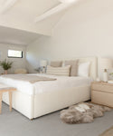 A bright, minimalist bedroom with a white bed, neutral pillows, light wood furniture, soft natural light from small windows, plants, simple table lamps, and a Hudson Jute Rug for a handwoven natural fiber touch.