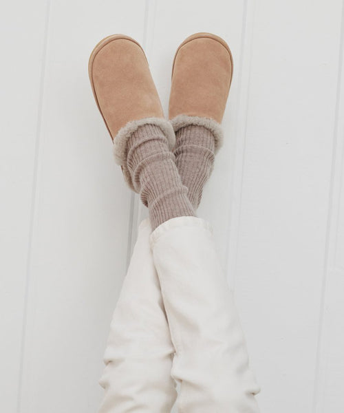 A person wearing Lodge Outdoor Slipper featuring a brown suede upper and cozy shearling lining, paired with white pants.
