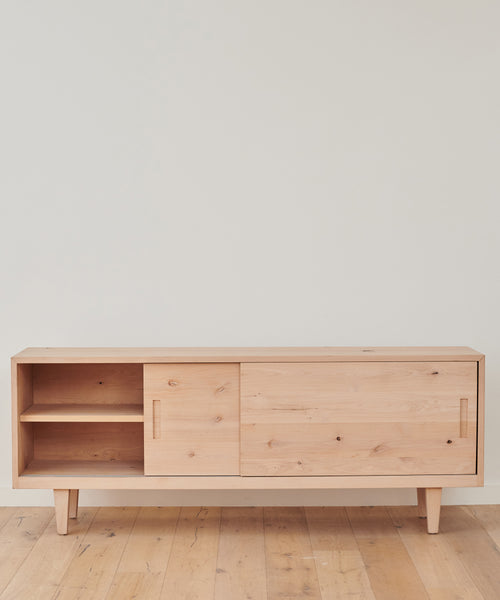 The Mandeville Credenza from Jenni Kayne Furniture, crafted from Knotty Alder with tapered legs, features an open shelf and two sliding doors for hidden storage. Its light wood finish complements the matching wood floor against a white wall.