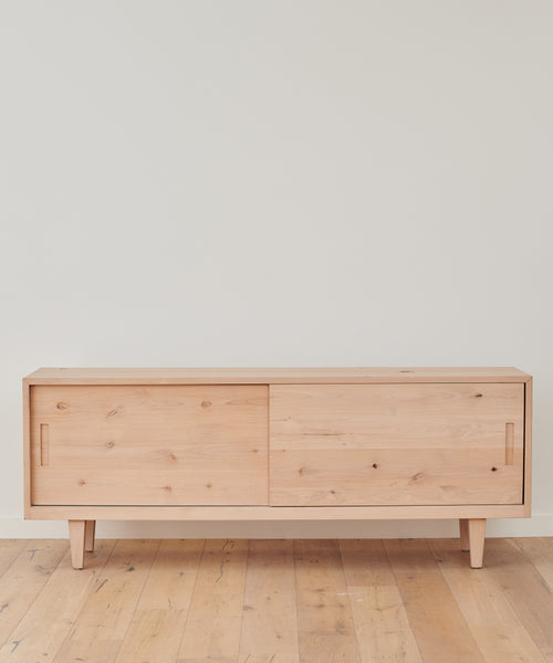 The minimalist Mandeville Credenza, crafted from knotty alder wood with two sliding doors and simple handles, stands against a plain white wall on a wooden floor.