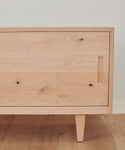 The Mandeville Credenza features a light wood finish, minimalist design, clean lines, a single door with a rectangular cutout handle, and angled legs. Its set on a wooden floor against a white wall in the Jenni Kayne Furniture style.
