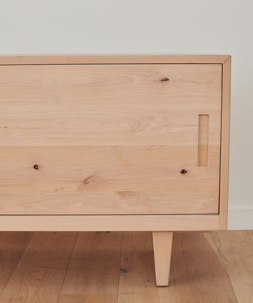 The Mandeville Credenza features a light wood finish, minimalist design, clean lines, a single door with a rectangular cutout handle, and angled legs. Its set on a wooden floor against a white wall in the Jenni Kayne Furniture style.