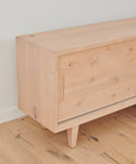 The Mandeville Credenza by Jenni Kayne Furniture, made of knotty alder with visible wood grain, has a handle cutout and angled legs. It sits on a light wood floor against a white wall.