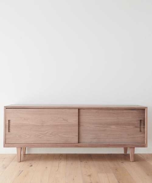 The Mandeville Credenza stands against a plain white wall on a light wood floor, featuring minimalist design with sliding doors and angled legs.