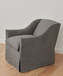 The handmade Miramar Chair, crafted in the USA, features gray Belgian linen with a fitted slipcover, curved arms, and a cushioned seat. It sits on a wooden floor against a plain white wall.