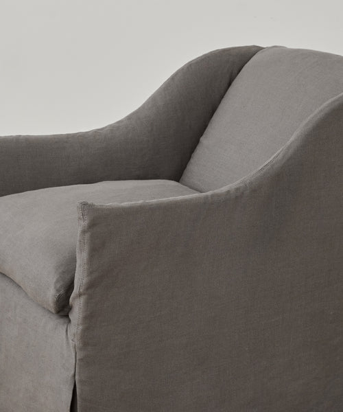 Close-up of the Miramar Chair Slipcover in soft, grey Belgian linen upholstery with rounded arms and a cushioned seat, set against a plain light background.