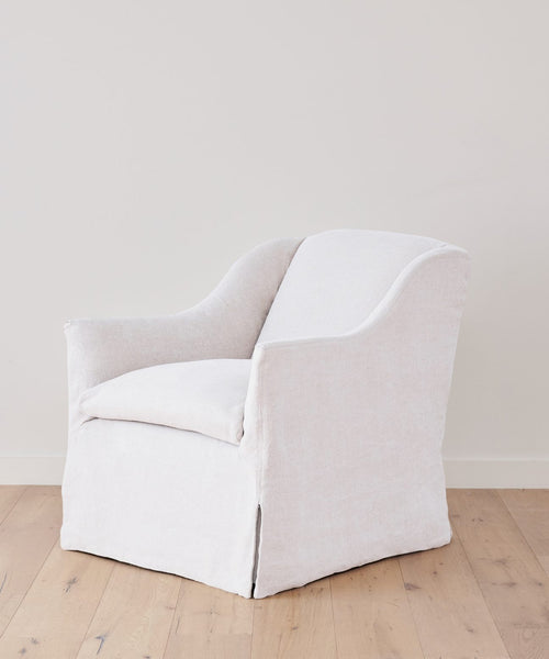 The Miramar Chair, upholstered in white linen with curved arms and a skirted base, stands on a light wood floor beside an off-white wall.