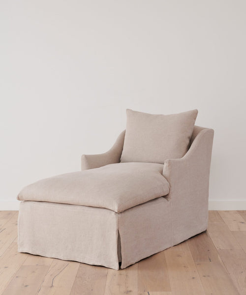 The Miramar Chaise Slipcover in Belgian linen covers a modern beige upholstered chaise lounge, set on a light wooden floor against a plain white wall.