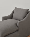 The Miramar Chaise Slipcover features soft gray Belgian linen upholstery, a thick cushion, and a matching pillow, creating a modern look against a plain light-colored wall.