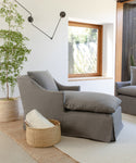 A cozy Miramar Chaise Slipcover in grey linen sits by a window next to a leafy potted plant. A woven basket with a rolled blanket is on the floor, and a modern black wall sconce hangs above.