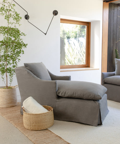 A cozy Miramar Chaise Slipcover in grey linen sits by a window next to a leafy potted plant. A woven basket with a rolled blanket is on the floor, and a modern black wall sconce hangs above.