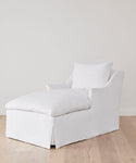 The Miramar Chaise Slipcover, featuring white linen and a matching back cushion, sits on a light wooden floor against a plain white wall.