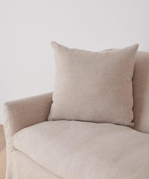 A light beige armchair with a square cushion, dressed in the Miramar Sofa Slipcover made of Belgian linen, sits on a wooden floor against a plain white wall.