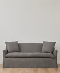 The Miramar Sofa Slipcover in minimalist gray linen, featuring two matching cushions, rests against a plain white wall on a light wooden floor.