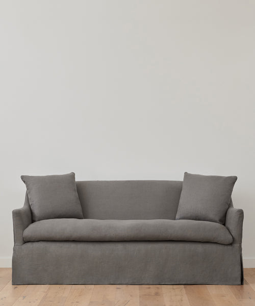 The Miramar Sofa Slipcover in minimalist gray linen, featuring two matching cushions, rests against a plain white wall on a light wooden floor.