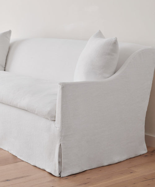 The Miramar Sofa, upholstered in light gray Belgian Libeco Linen with two matching cushions, showcases a modern design with soft curves and clean lines, set against an off-white wall on a wooden floor.