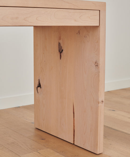 Close-up of a Newport Desk leg crafted from Knotty Alder, showcasing natural knots and cracks, set on a light wood floor against a white wall.