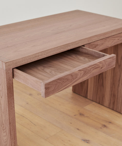 A close-up of the Newport Desk showcases its natural wood grain and smooth finish, with a partially open drawer. This solid wood desk stands on a wooden floor against a light-colored background.