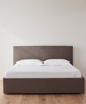 The Pacific Bed features a gray upholstered headboard and frame, dressed in white Belgian linen sheets and two white pillows. It stands on a light wooden floor against a plain white wall.