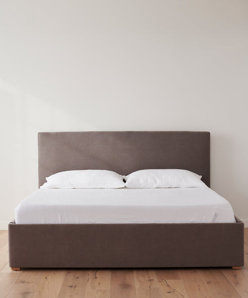 The Pacific Bed features a gray upholstered headboard and frame, dressed in white Belgian linen sheets and two white pillows. It stands on a light wooden floor against a plain white wall.