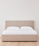 The Pacific Bed, featuring modern beige upholstery and a tall Belgian linen headboard, is styled with white bedding and two pillows in a minimal room with light wood floors and a plain white wall.