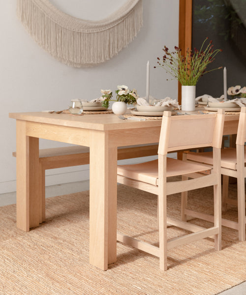 A light wooden dining table set with neutral tableware, white candles, and fresh flowers is surrounded by light wood chairs. A Rug Swatch and woven wall hanging add cozy texture to this minimalist space.