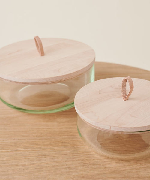 Two Stowe Eco Glass Bowls made from recycled glass, featuring light FSC-certified wooden lids and leather loop handles, are placed on a wooden surface against a light background.