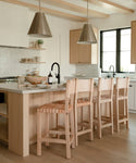 A modern kitchen features light wood Knotty Alder cabinets, a white island with three Tahoe Counter Stools, two metal pendant lights, open shelves displaying dishes, and a black faucet by the window.