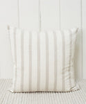 The Ojai Pillow, featuring beige and light gray vertical stripes, is handwoven and rests against a white paneled wall on a light, textured carpet.