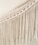 Close-up of the Woven Wall Hanging featuring cream-colored macramé cotton with twisted, knotted rope designs at the top and long, loose fringes cascading below.
