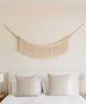 A minimal bedroom features a white headboard, two beige pillows on a bed with white bedding, and the Woven Wall Hanging in cream above the bed, adding subtle handmade decor to the plain light-colored wall.
