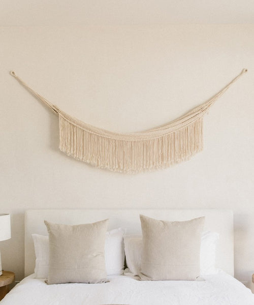 A minimal bedroom features a white headboard, two beige pillows on a bed with white bedding, and the Woven Wall Hanging in cream above the bed, adding subtle handmade decor to the plain light-colored wall.